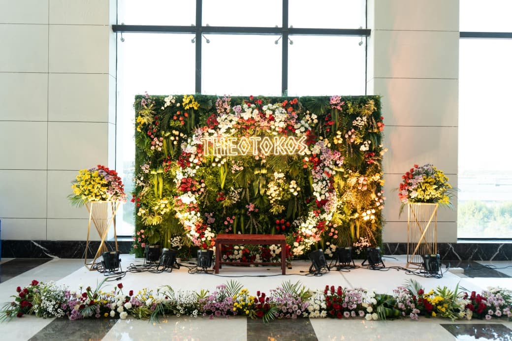 Floral wall with a glowing Theotokos sign, lined with fresh flowers below